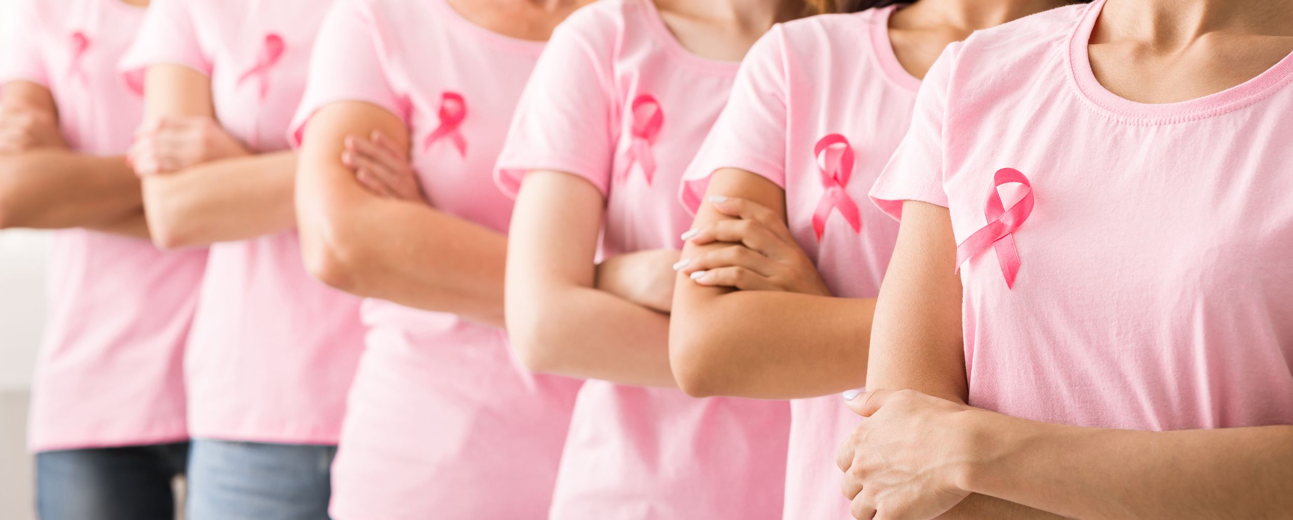 A row of women standing side by side with arms crossed, all wearing pink shirts with pink ribbons pinned on them, symbolizing breast cancer awareness and solidarity.