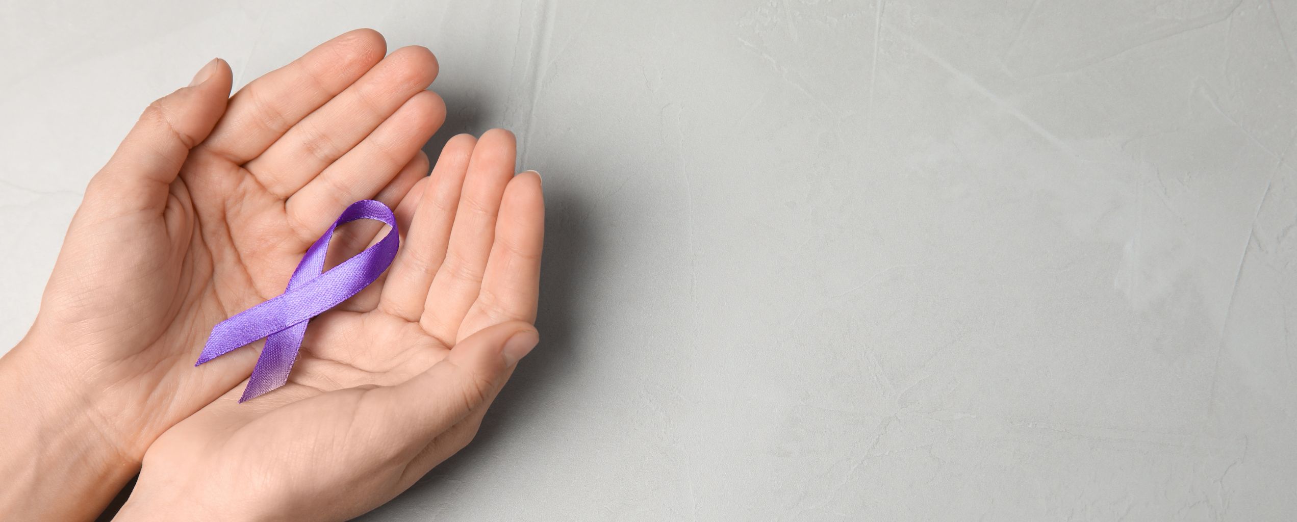 Two hands gently cupping a purple awareness ribbon against a light gray background, symbolizing domestic violence awareness and support.